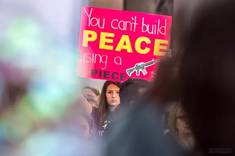 Image: A girl holds a poster that reads "You can't build peace using a piece" with a photo of a gun.