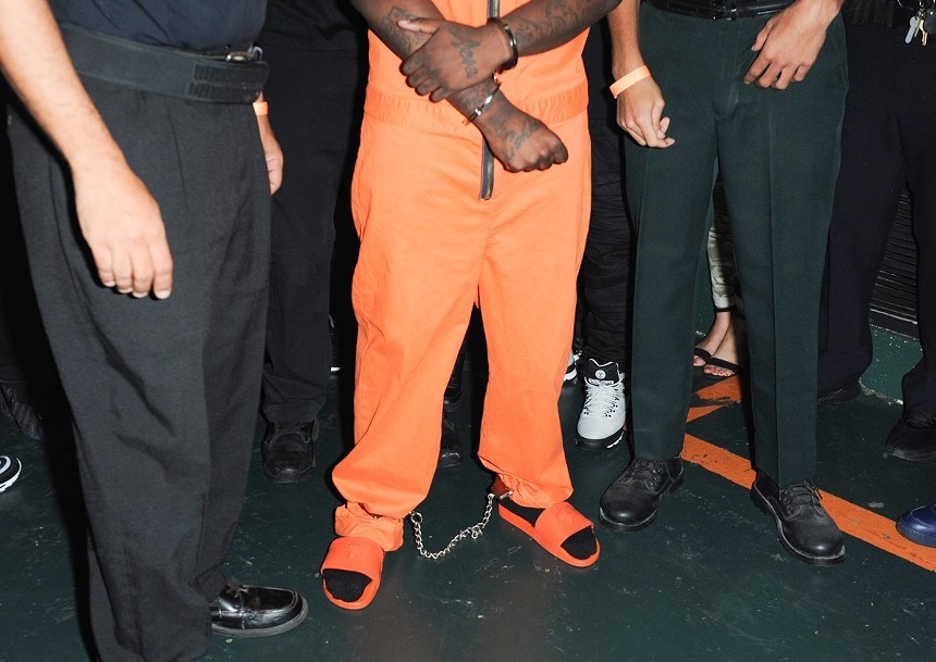 Kodak Black in a posed color shot, dressed in an orange prison jumpsuit, standing between two white men in police gear